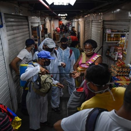Un trabajador municipal que llevaba un traje protector rocía desinfectante en las manos de un cliente en un mercado en el barrio de Petare después de que el gobierno alivió un bloqueo nacional impuesto como medida preventiva contra el coronavirus COVID-19, en Caracas, el 13 de julio de 2020. (Foto de Federico PARRA / AFP)